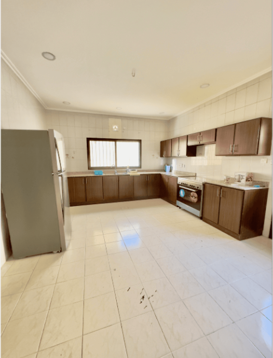 Spacious kitchen with white tiled walls and floor, dark wooden cabinets, stainless steel refrigerator, oven, and a window.